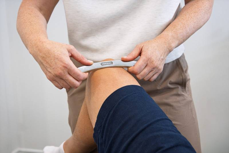 A chiropractor using a stainless-steel Graston instrument above a patient’s knee