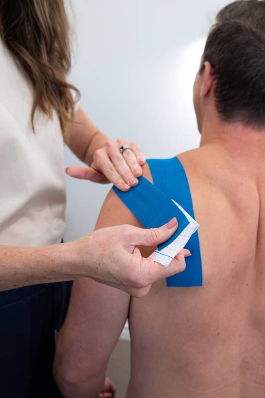 A chiropractor applying blue kinesiology tape across a patient’s shoulder